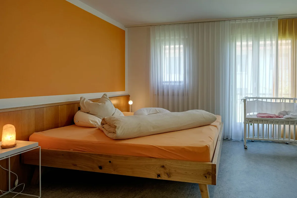Bild eines Betts mit Kissen und Decke und einem Nachttisch, vornedran ein Babybett.